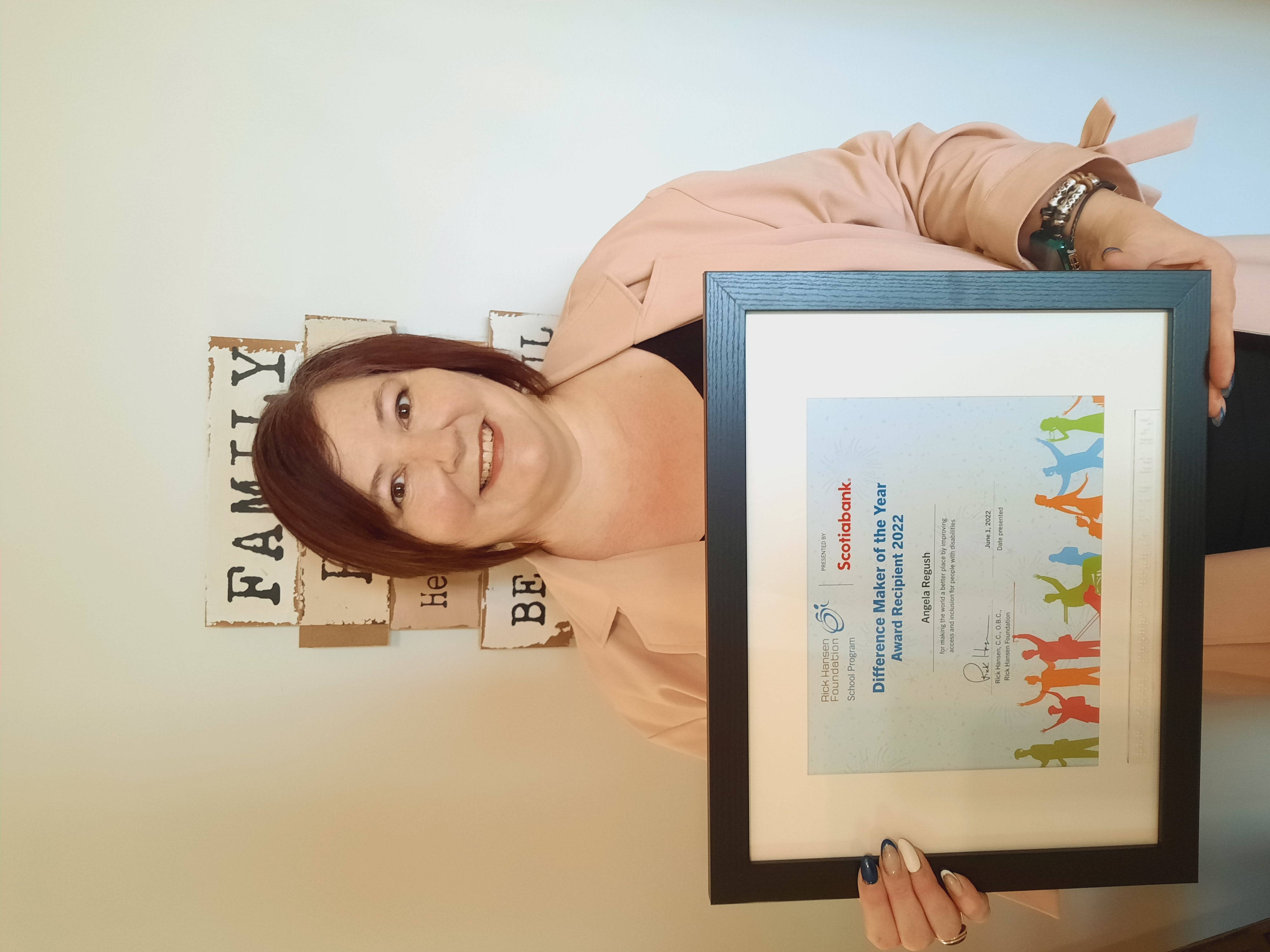 Woman with short red hair holding a framed Different Maker award certificate. She is wearing a pink blazer and standing in front of a sign that says family. 